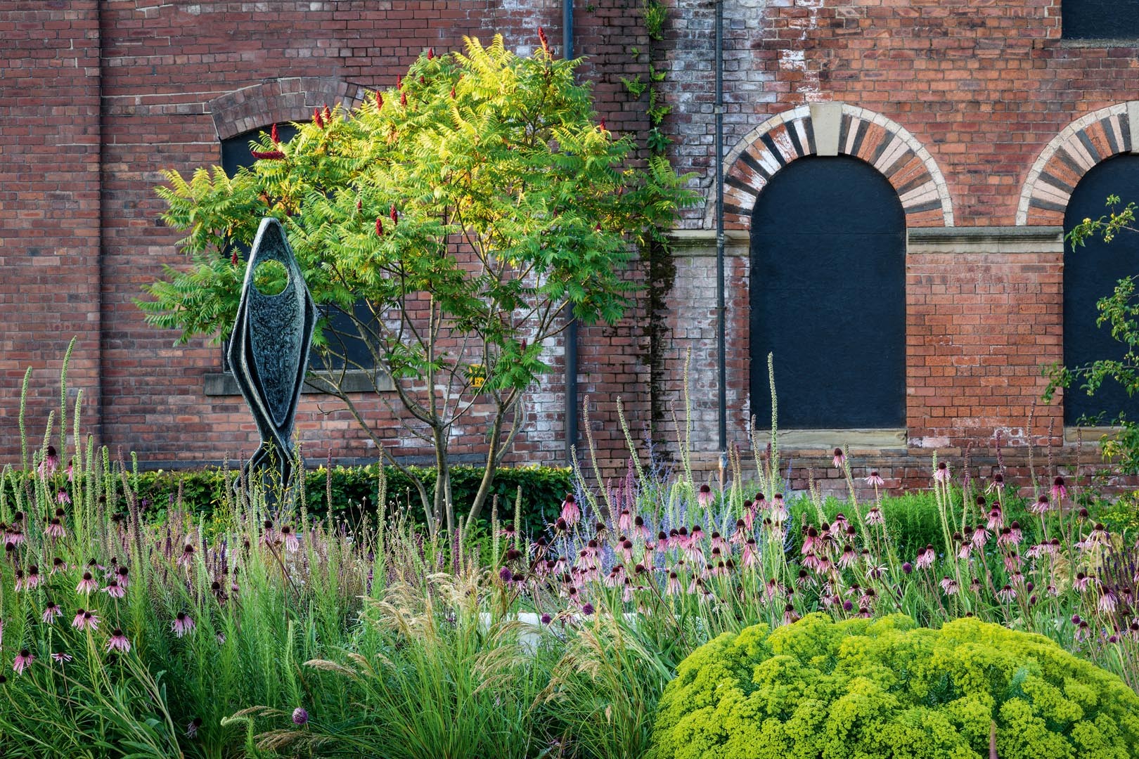 The red brick walls of the old Rutland Mills highlight the intensity of the summer green foliage of the Fagus sylvatica hedge and the foliage of the Rhus typhina that shades the sculpture Ascending Form (Gloria). Across the path Liatris pycnostachya adds height to a mix of Stipa calamagrostis, Echinacea pallida and Euphorbia seguieriana.