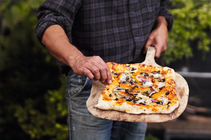 A pizza that has been cooked over a campfire has been placed on a wooded utensil and is ready to serve